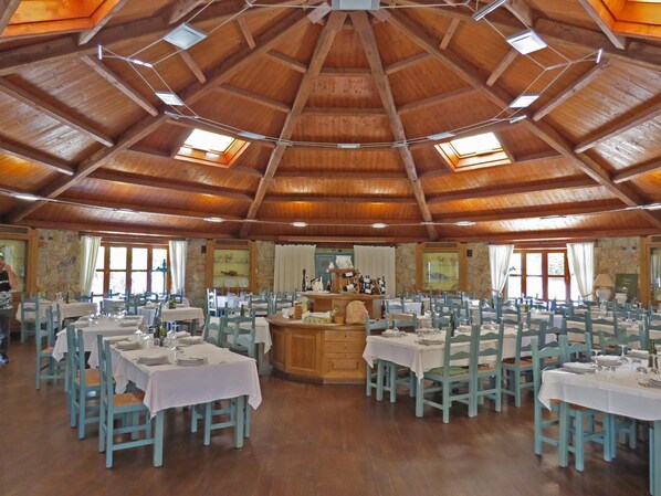 A large dining hall with multiple tables and chairs, a central bar area, and a high wooden ceiling with skylights.