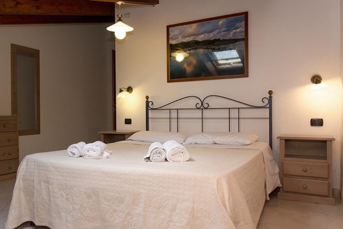 A neatly made bed with rolled towels, a bedside table, a nightstand, a wall-mounted lamp, and a framed picture on the wall.