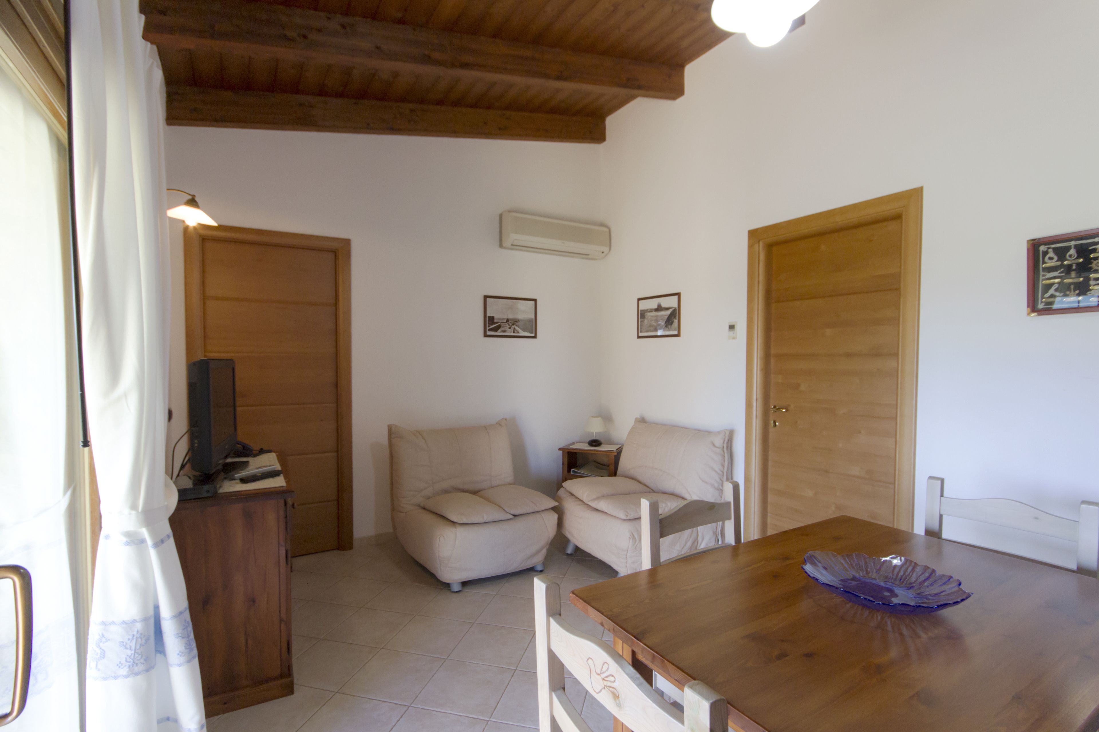 A room with a wooden table, two beige armchairs, a wooden cabinet, and a television on a stand.