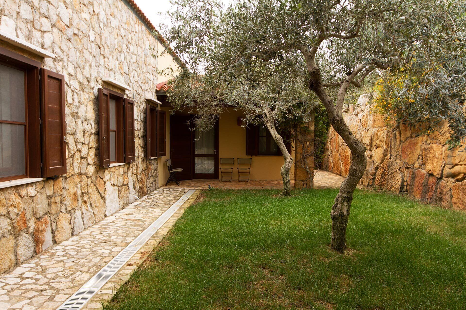 A stone building with a tiled pathway leading to a door, a small tree, and a grassy area.
