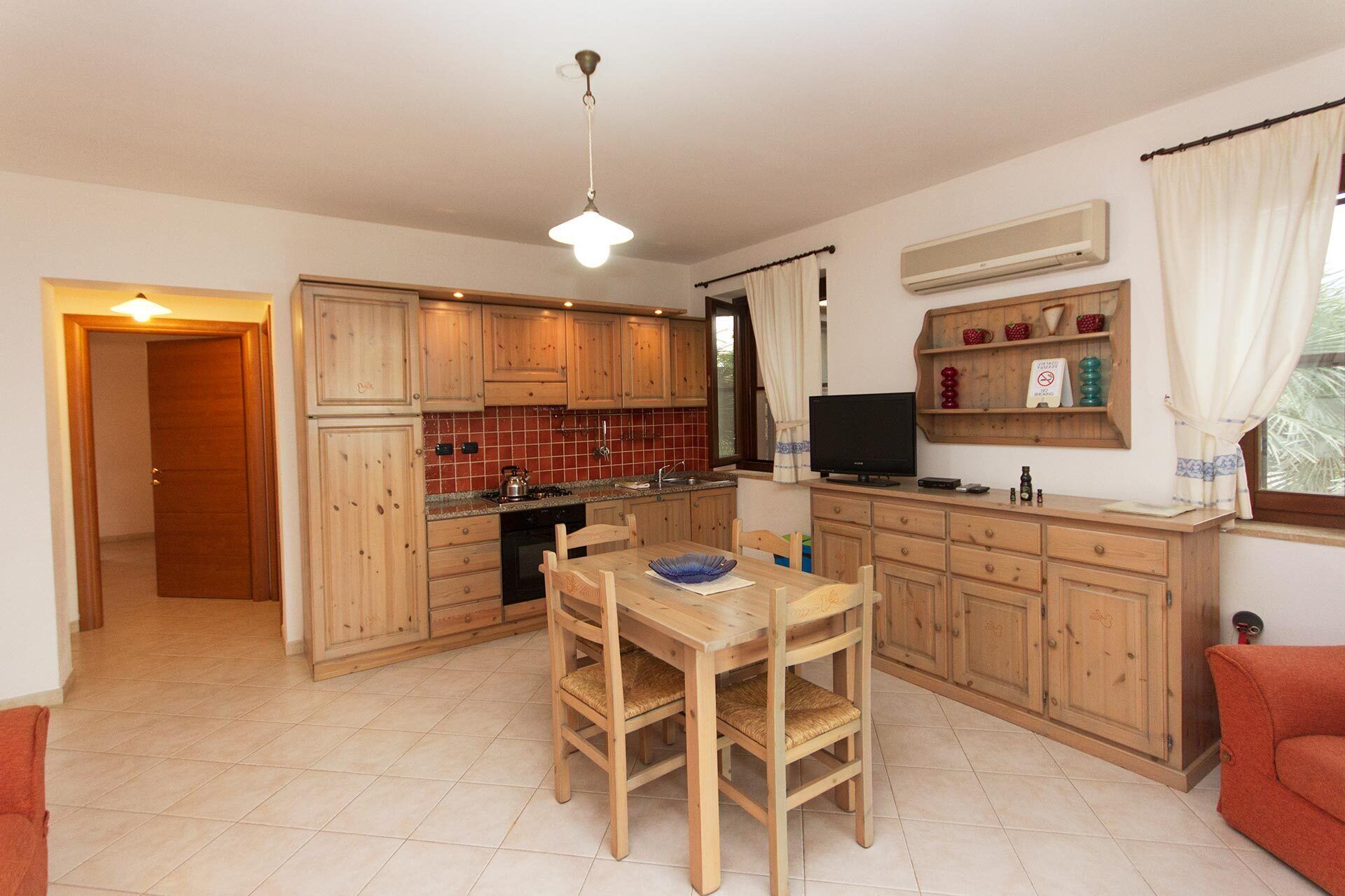 A compact kitchen with wooden cabinets, a dining table with chairs, a TV, and a small shelf with decorative items.
