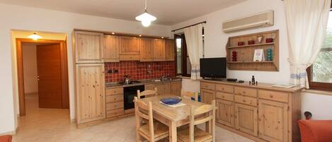 A compact kitchen with wooden cabinets, a dining table with chairs, a TV, and a small shelf with decorative items.