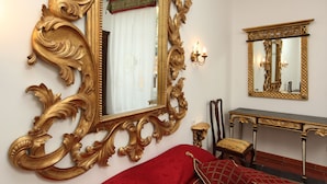 A room with a large ornate gold-framed mirror, a red upholstered bed, a wooden console table, and a small chair with a cushion. - Navona Governo Vecchio (Rome)