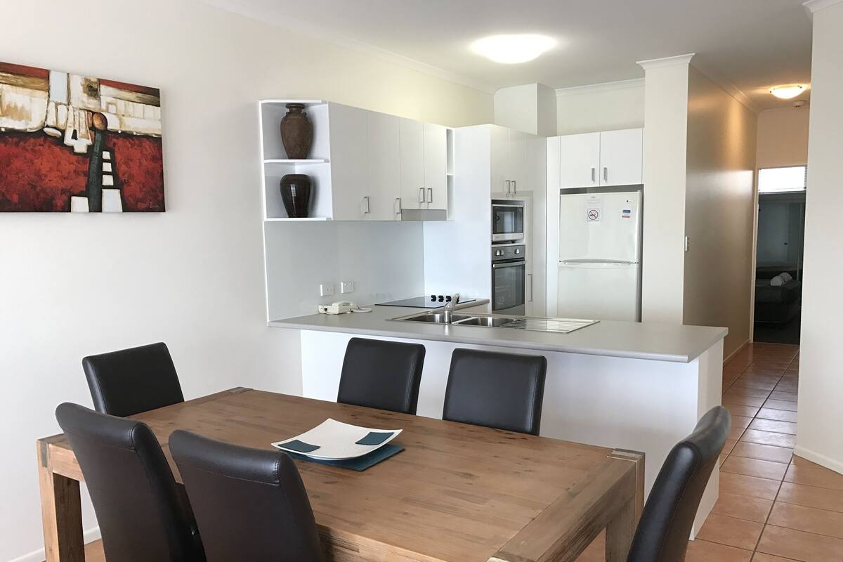 Une cuisine moderne avec des meubles blancs, un four encastré et un réfrigérateur. On y trouve une table à manger avec quatre chaises et un vase accroché au mur.