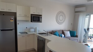 Eine moderne Küche mit Kühlschrank, Mikrowelle, Toaster und Spüle. Es gibt einen Essbereich mit einem weißen Tisch und braunen Hocker. Im Hintergrund steht ein Sofa mit blau-weißen Kissen.