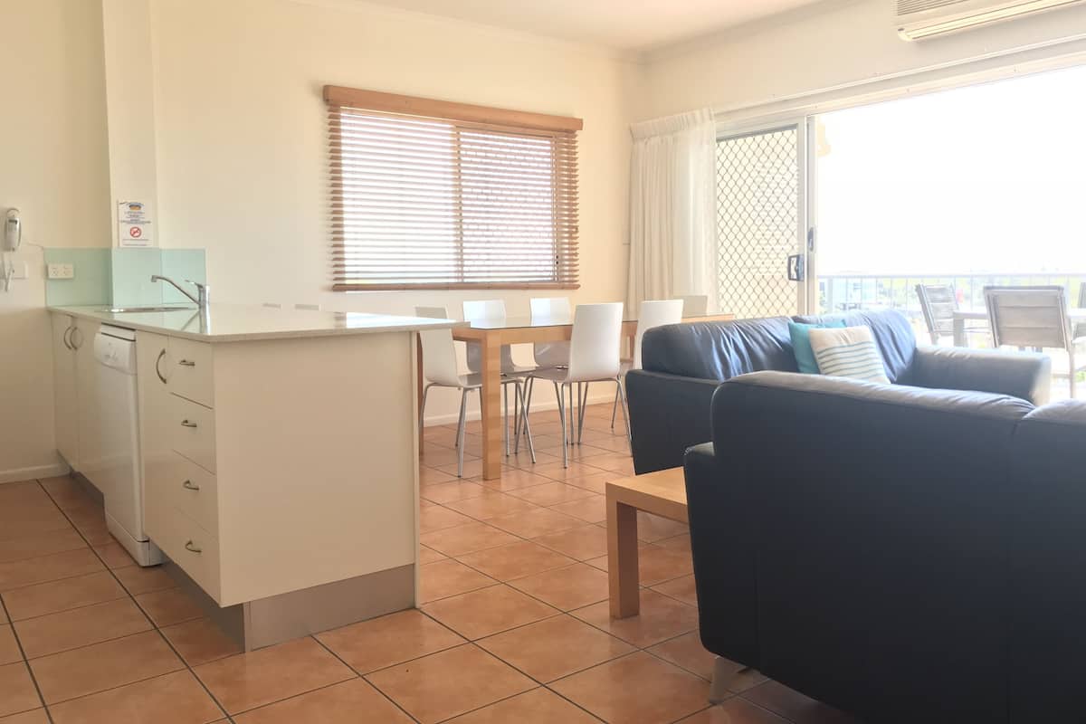 Un salon moderne comprenant un canapé, une table à manger avec des chaises, un coin cuisine avec un plan de travail et un évier, ainsi qu’un balcon avec vue.