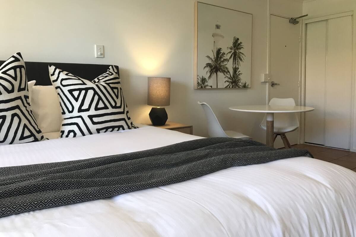 Une chambre d’hôtel avec un lit, une table de chevet avec une lampe, une chaise, une petite table et une porte.