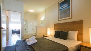 Ein Hotelzimmer mit Bett, Nachttisch, einer Nachttischlampe, einem Badezimmer mit Dusche und einem Bild mit einer Strandansicht.