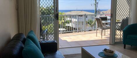 A living room with a leather sofa, a coffee table, and a view of a balcony with outdoor furniture and a building in the background.