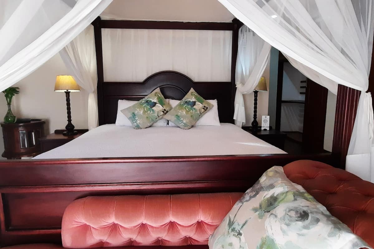 Un dormitorio con cama con dosel, ropa de cama blanca y almohadas florales. Hay una mesita de noche con una lámpara y un jarrón de flores. Un banco tapizado en rojo se encuentra frente a la cama.