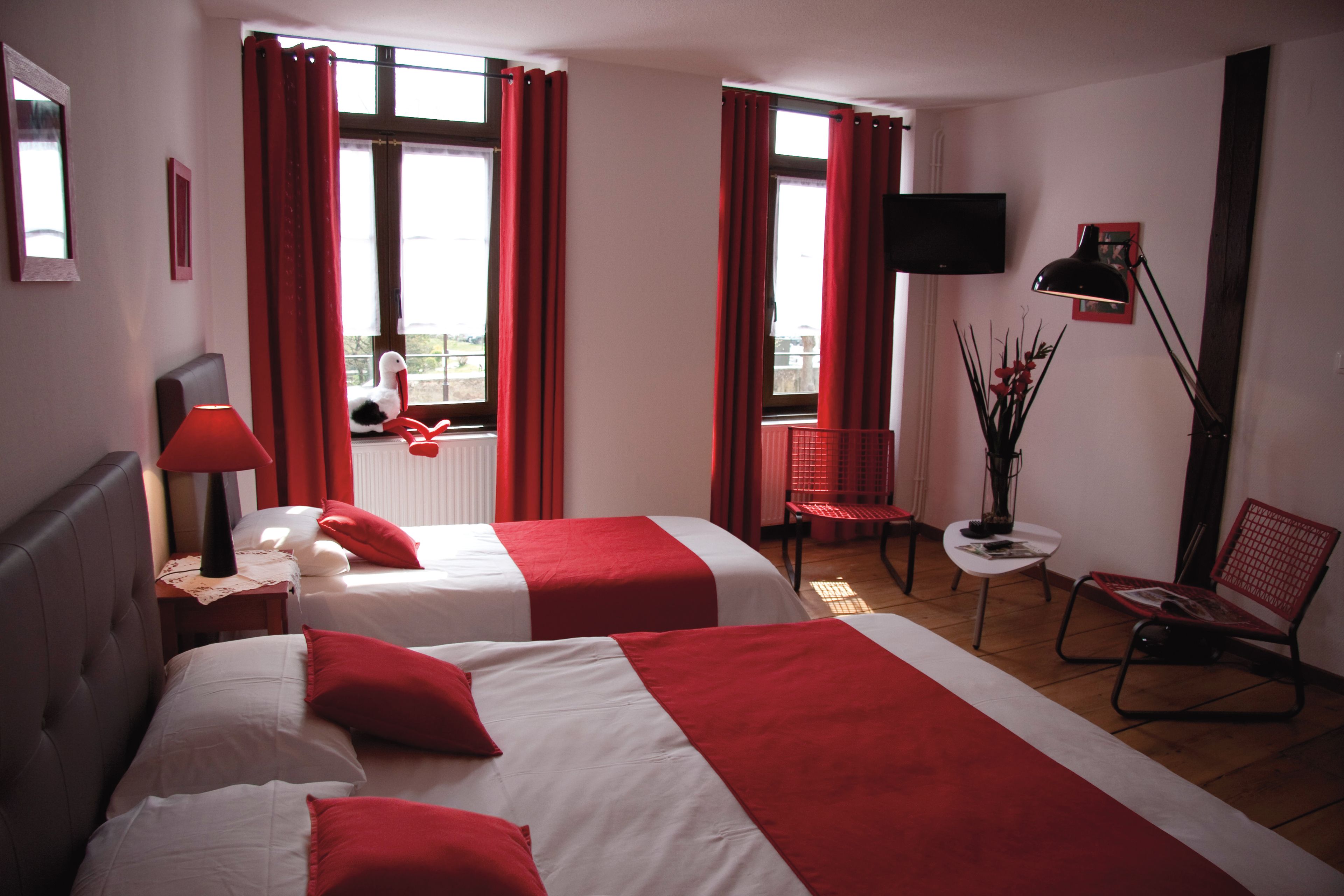 Habitación de hotel con dos camas, ropa de cama roja y blanca, un televisor, un escritorio con lámpara, una silla y una ventana con cortinas.