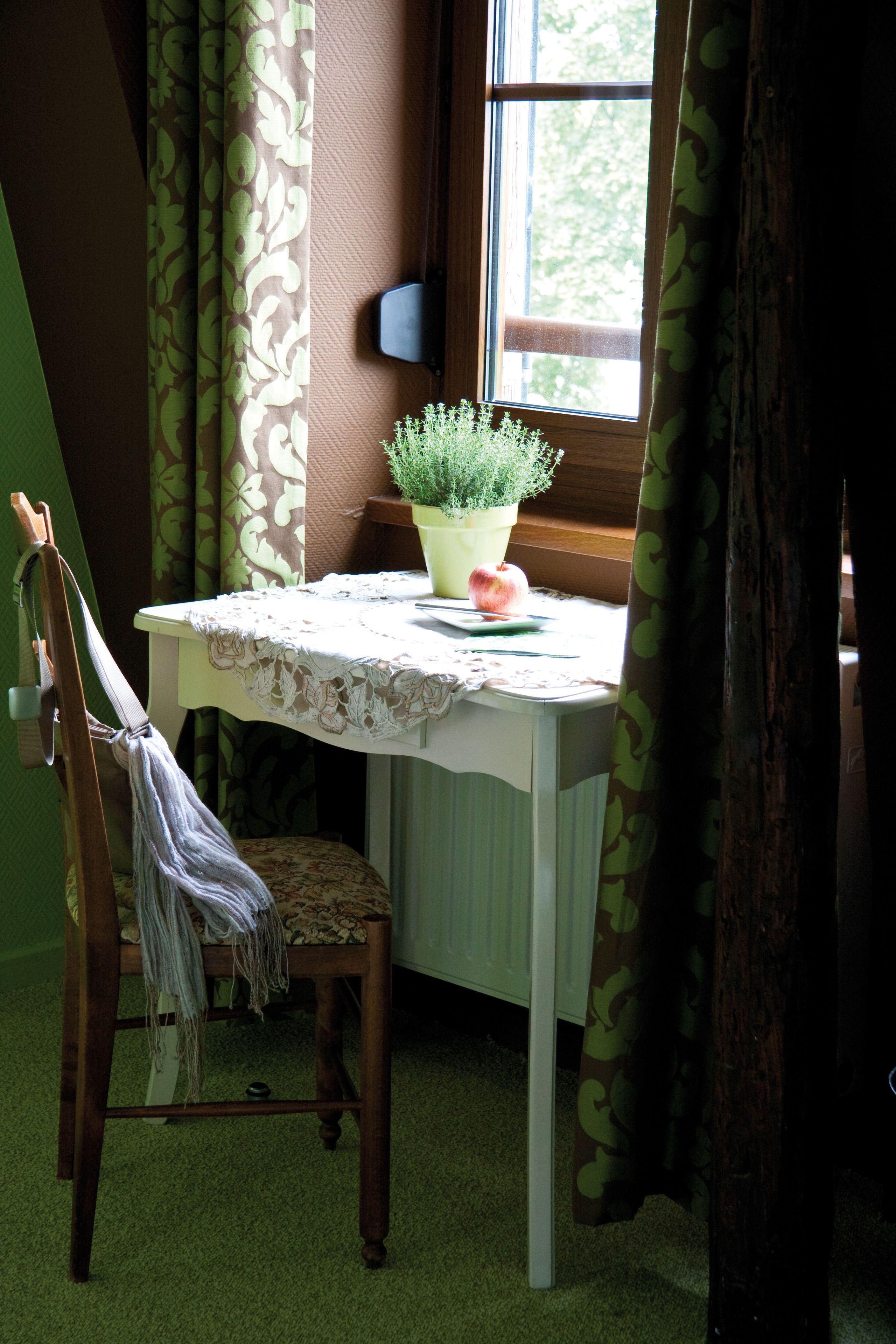 A room with a table by a window, a chair, a potted plant, and a small table with a lace tablecloth.