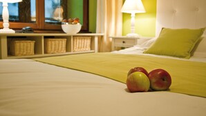 Cama bem arrumada com manta verde, duas maçãs sobre a cama, cabeceira branca e mesa de cabeceira com abajur e uma cesta de frutas.