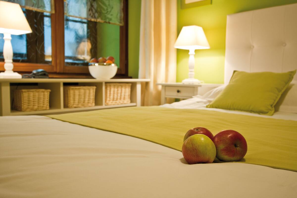 A neatly made bed with a green throw blanket, two apples on the bed, a white headboard, and a nightstand with a lamp and a bowl of fruit.