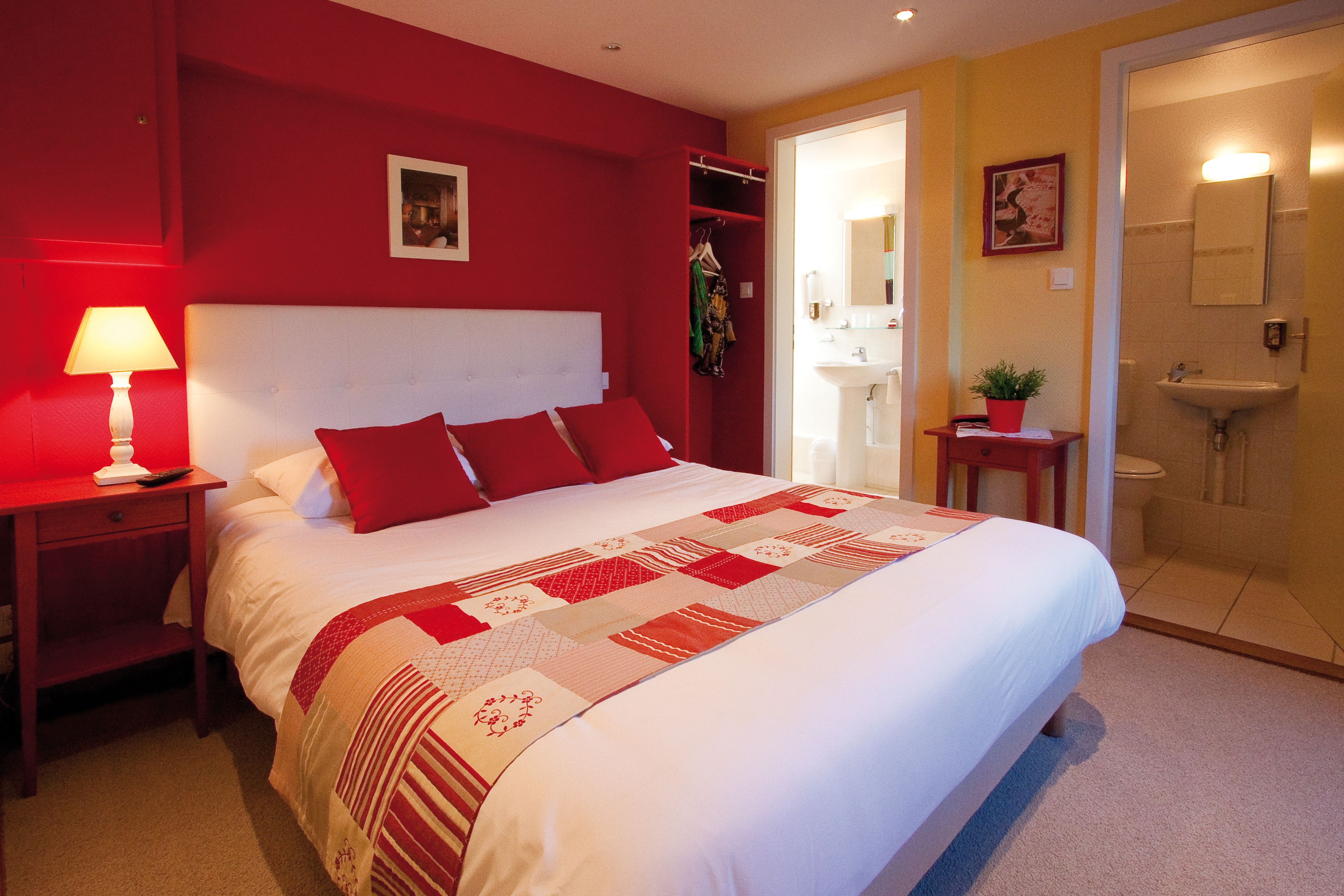 Un dormitorio con una cama grande, una mesita de noche, una pared roja, una puerta a un baño y una pequeña mesa con una planta.