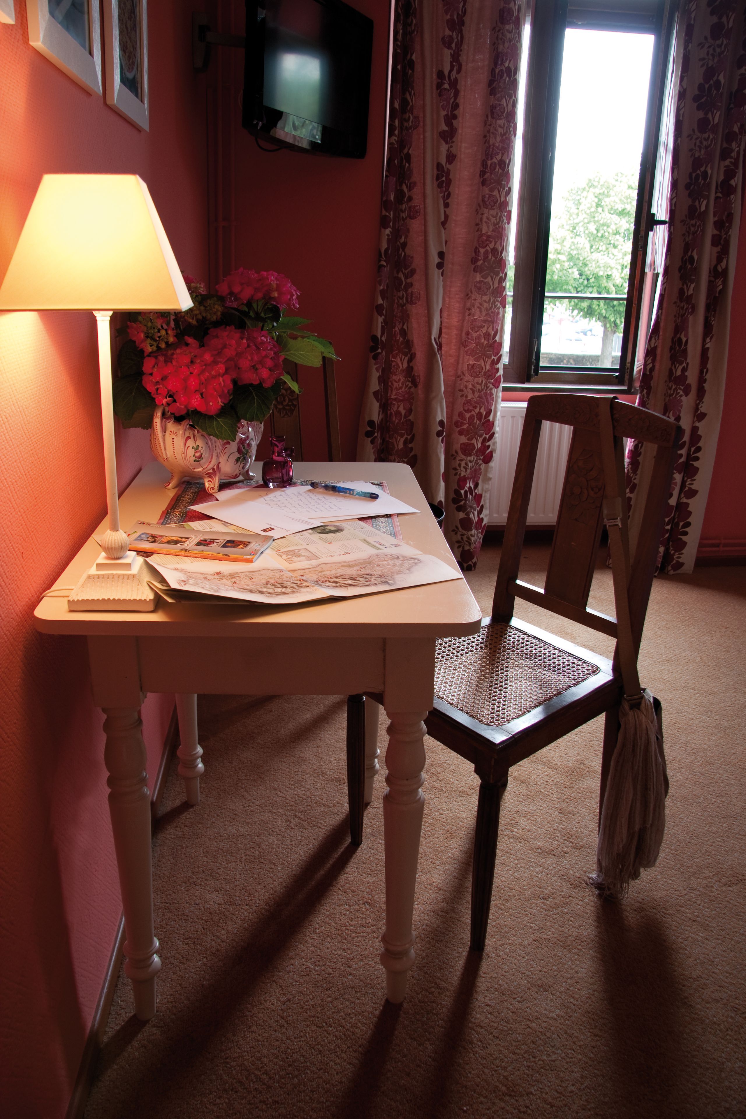 Una habitación con una mesa, una lámpara, una silla, un florero con flores y un televisor en la pared.