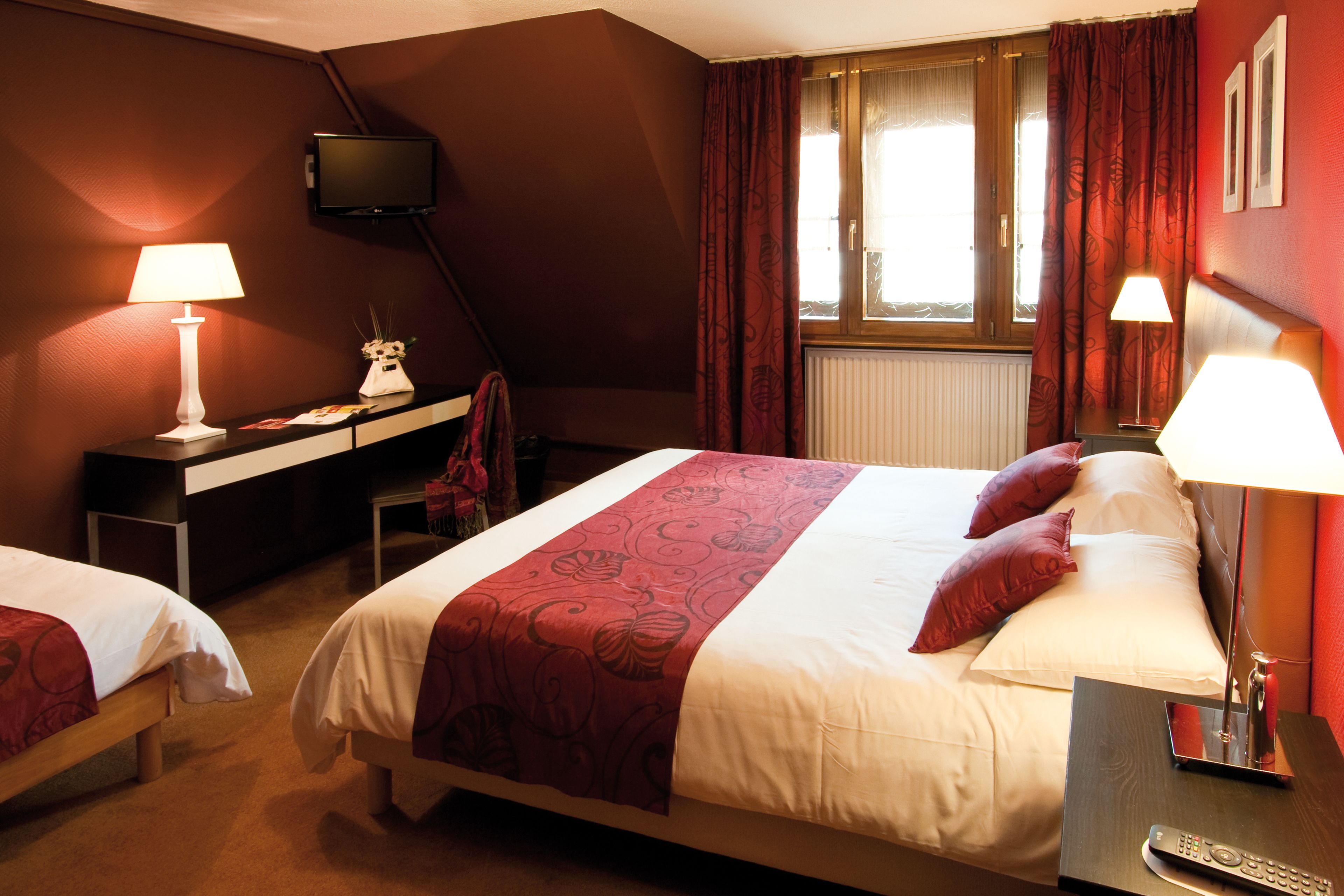 A hotel room with two beds, a desk with a lamp, a TV on the wall, and a window with curtains.