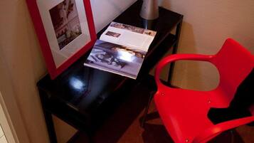 Een hoek van een kamer met een bureau, een rode stoel, een ingelijst beeld en een lamp.