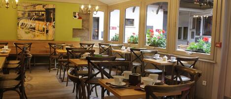 A restaurant with wooden tables and chairs, a green wall with a painting, and large windows with flowers.