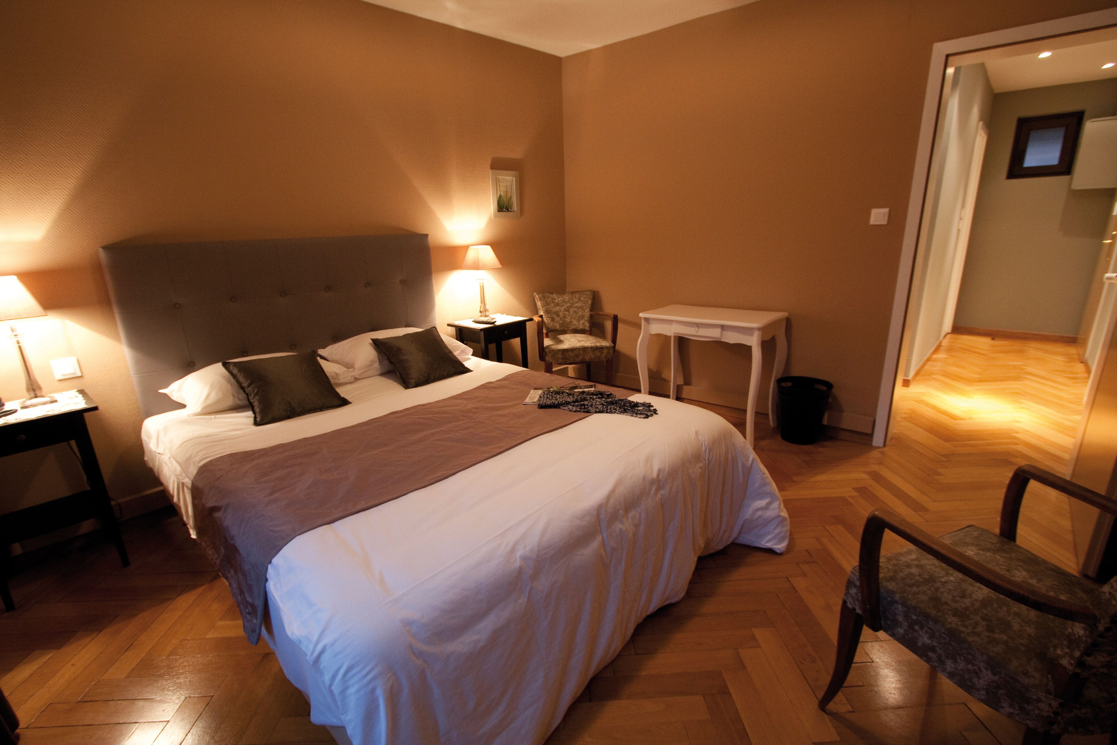 A hotel room with a large bed, two bedside tables with lamps, a chair, a small table, and a wooden floor.