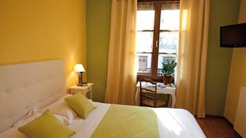 Una camera d'albergo con un letto, un comodino con una lampada, una sedia, un tavolo con una pianta e una TV montata a parete.