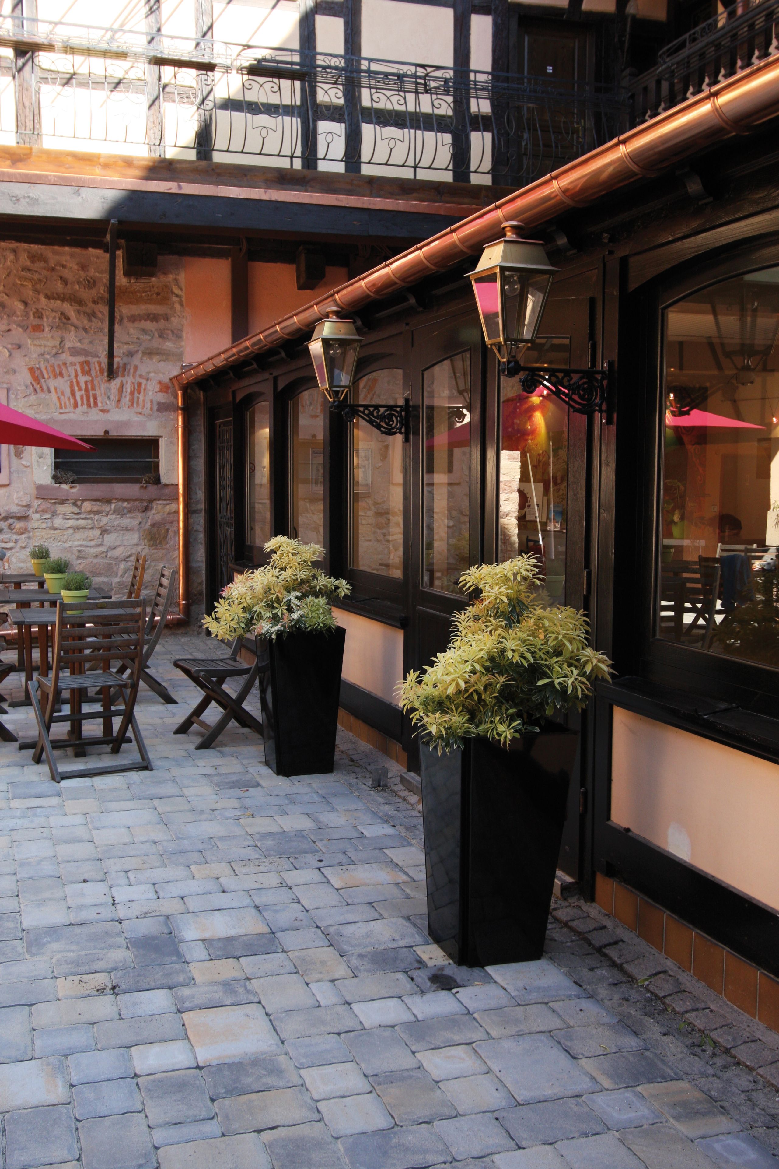 An outdoor seating area with tables and chairs, potted plants, and a building facade with large windows and a balcony.