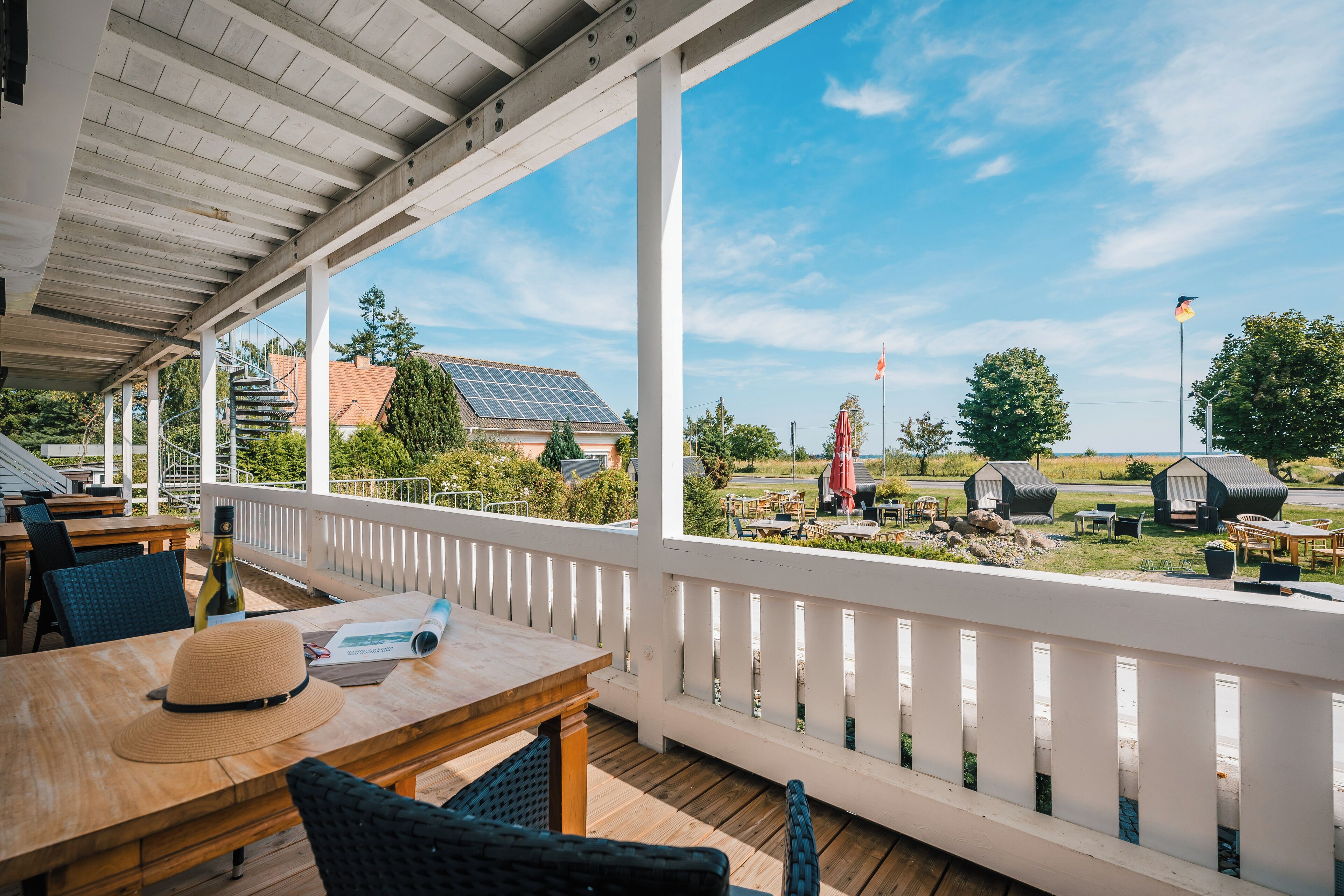A covered outdoor area with wooden tables and chairs, a white railing, and a view of the surrounding landscape including solar panels and outdoor furniture.