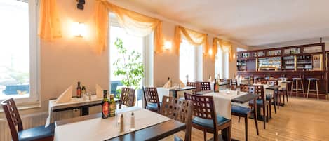 A restaurant with wooden tables and chairs, a bar area, and large windows with curtains.