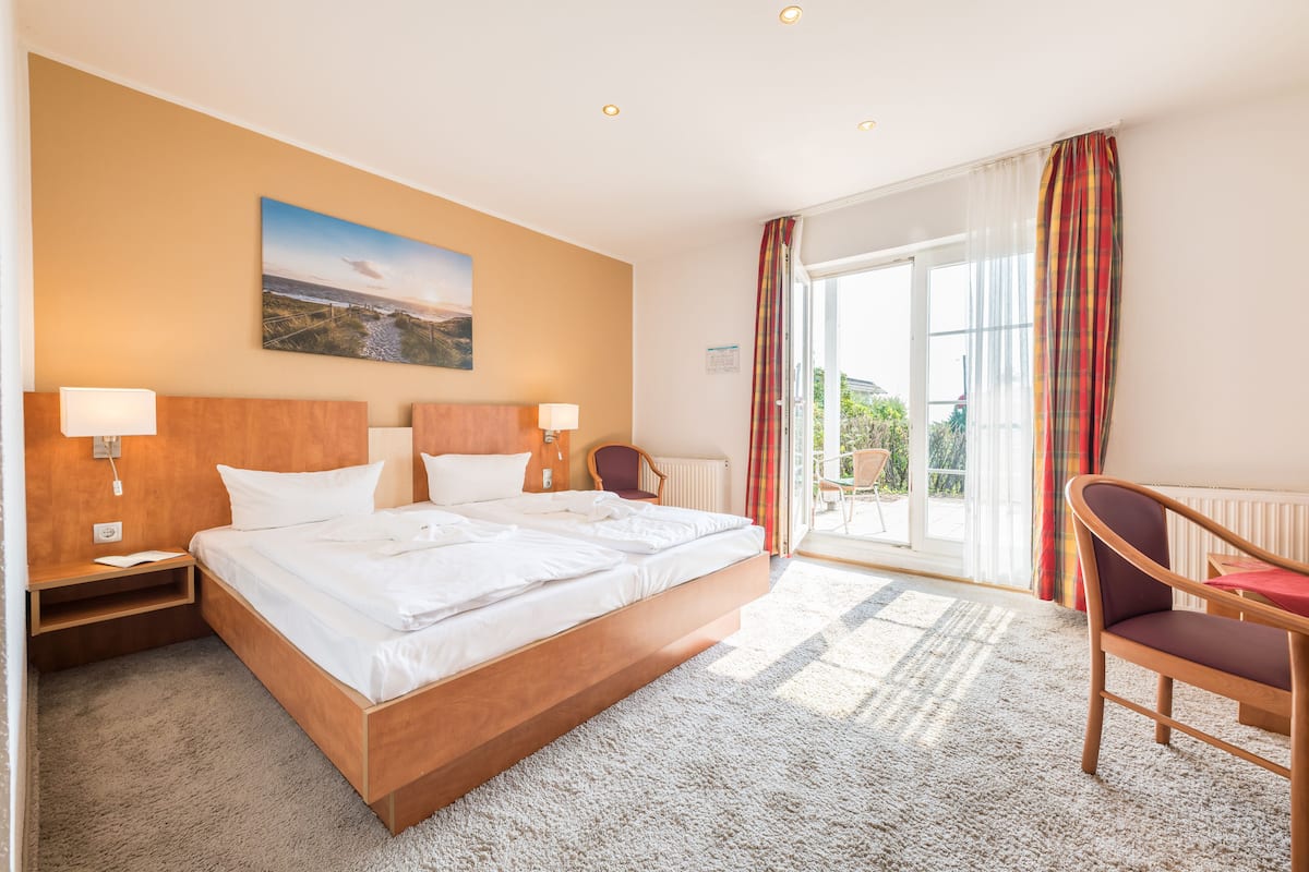 A modern hotel room with a large bed, two bedside tables with lamps, a chair, and a view of the outdoors through a sliding glass door.