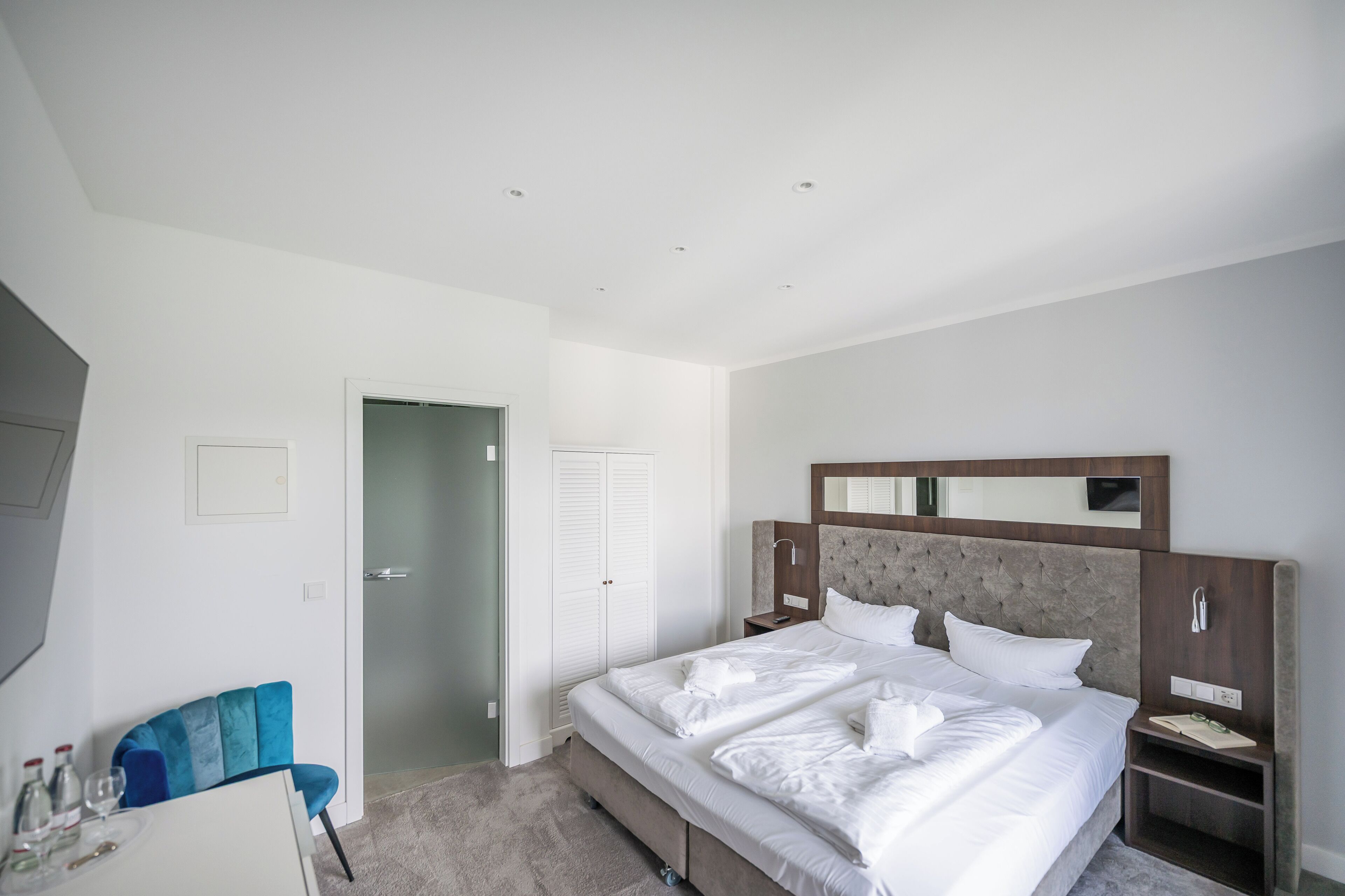 A modern hotel room with a large bed, a bedside table, a nightstand, a mirror, and a seating area with a chair and table.