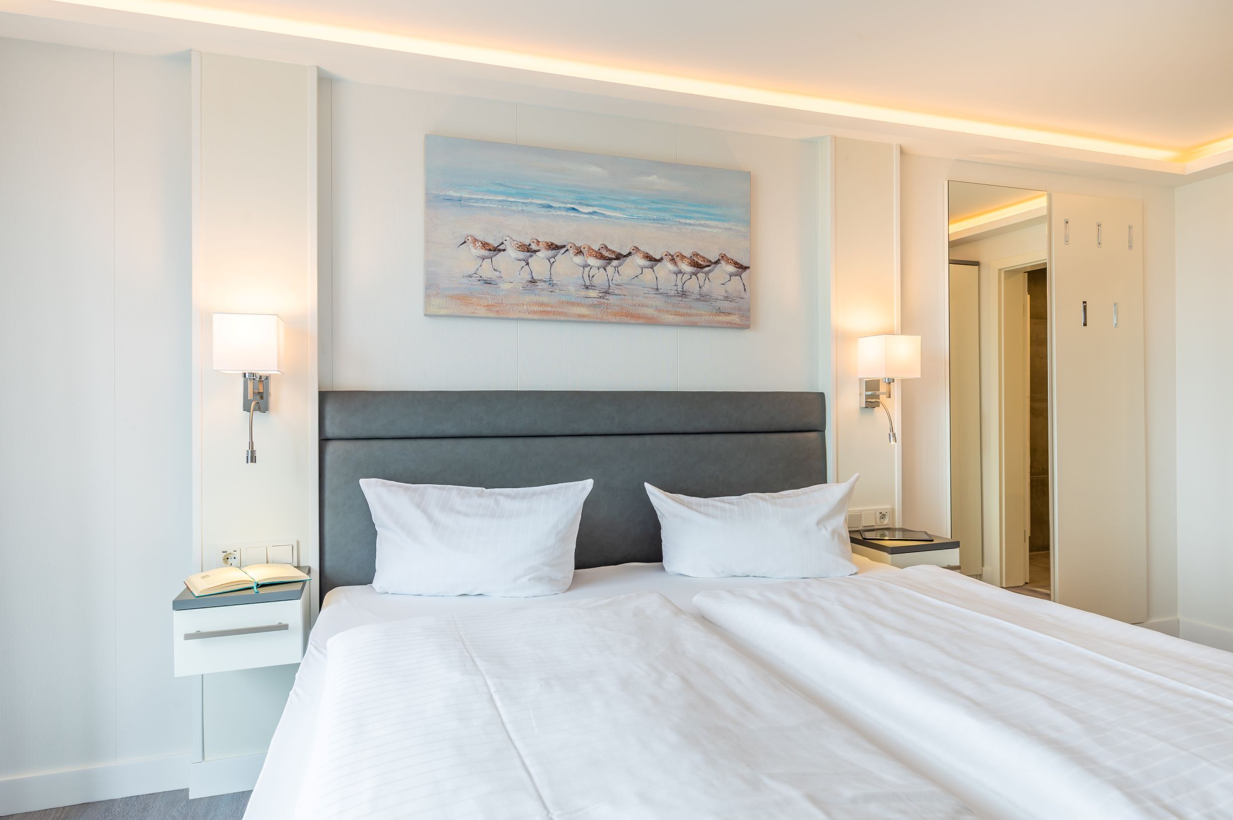 A hotel room with a large bed, two bedside tables, a nightstand, a painting of birds on the wall, and a door leading to another room.