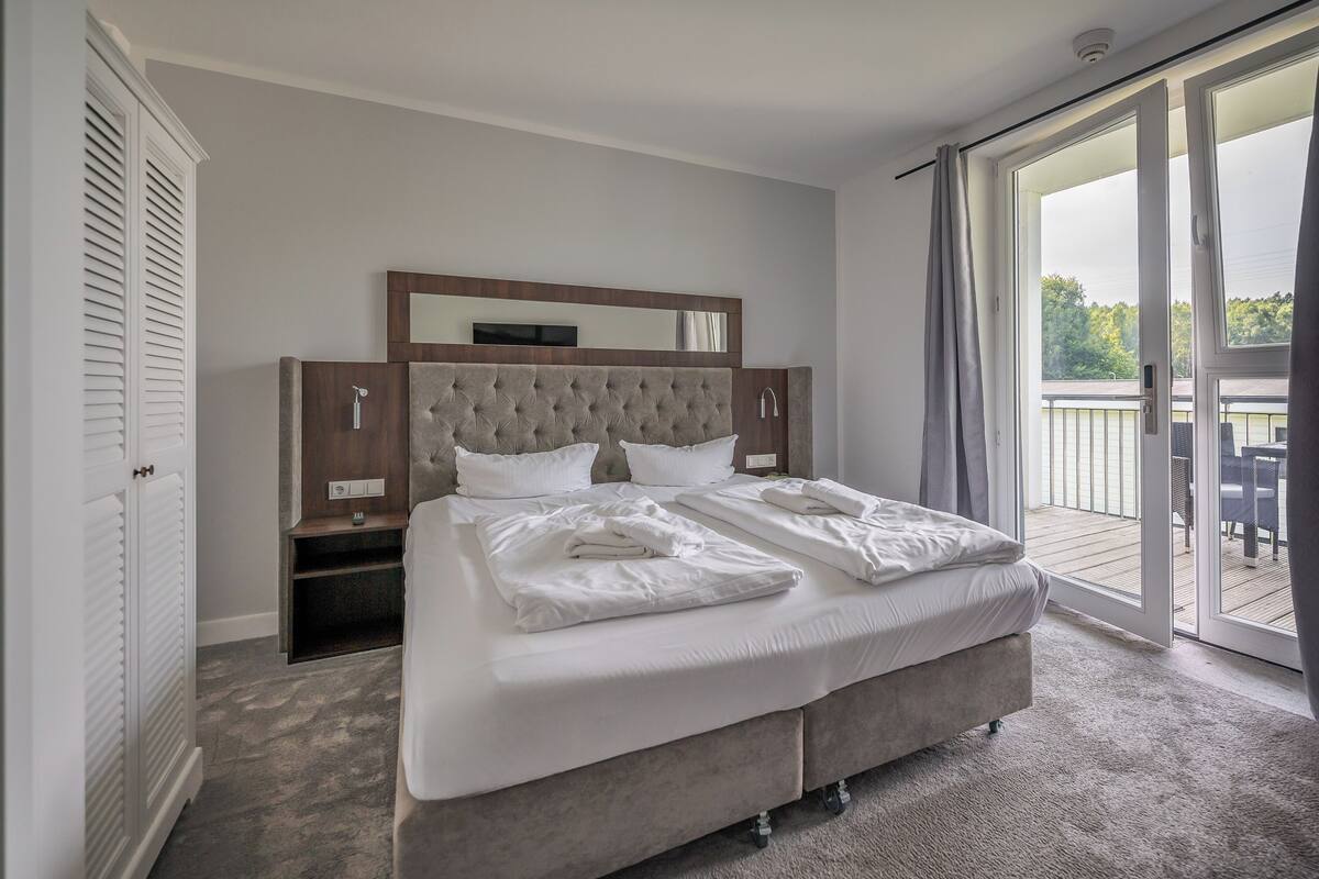 A bedroom with a large bed, a TV, a mirror, and a sliding glass door to a balcony.