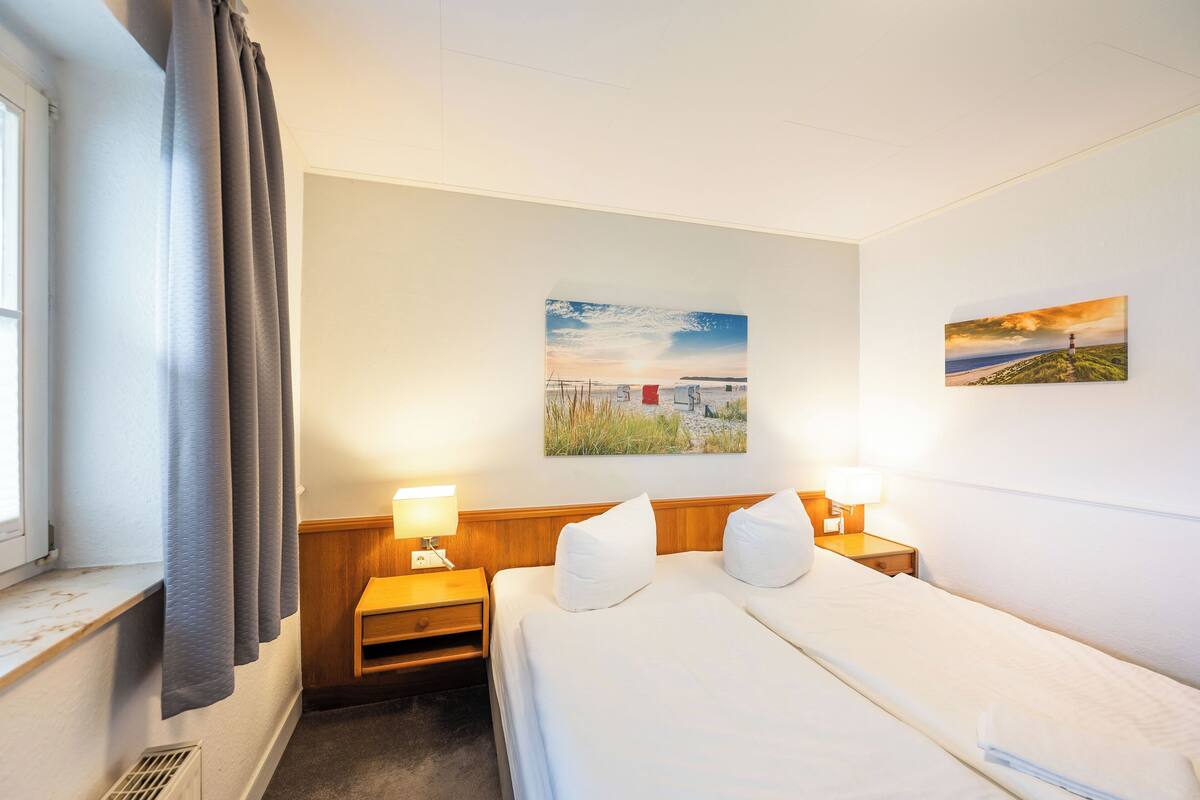 A hotel room with a bed, two bedside tables, a window with curtains, and two wall-mounted paintings.