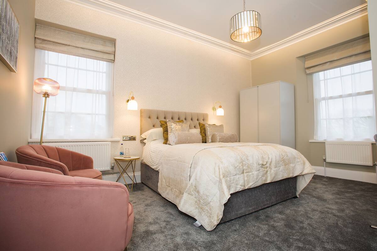 A bedroom with a large bed, two bedside lamps, a radiator, and a window with blinds.