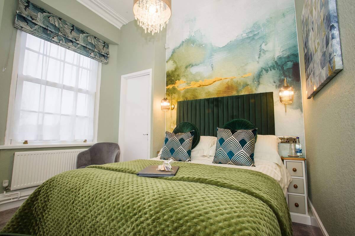 A bedroom with a green quilted bedspread, a headboard with a painting, a bedside table with a lamp, and a window with curtains.