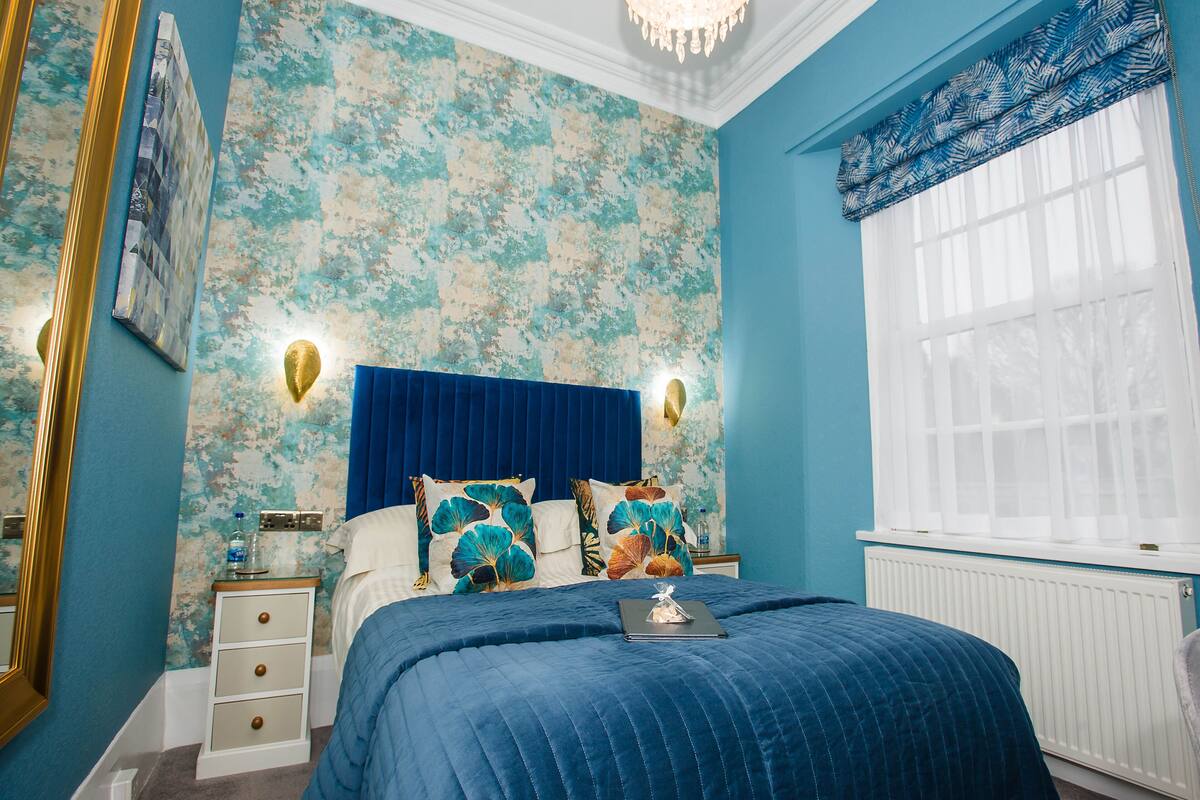 A bedroom with a blue bedspread, a floral wallpaper, a white radiator, and a window with a blue valance.
