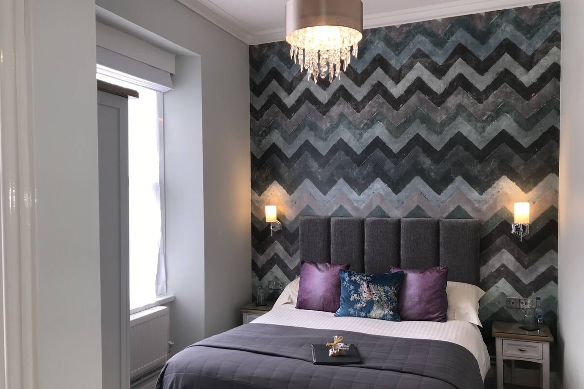 A modern bedroom with a chevron-patterned wallpaper, a large bed with multiple pillows, bedside lamps, and a chandelier.