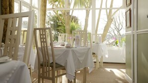 Un espace repas avec des nappes blanches, des chaises en bois et une vue sur les palmiers, visible à travers de grandes fenêtres.