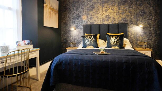 Une chambre avec une couvre-lit bleu foncé, une tête de lit agrémentée de deux coussins décoratifs, une table de chevet avec une lampe et un bureau avec une chaise.