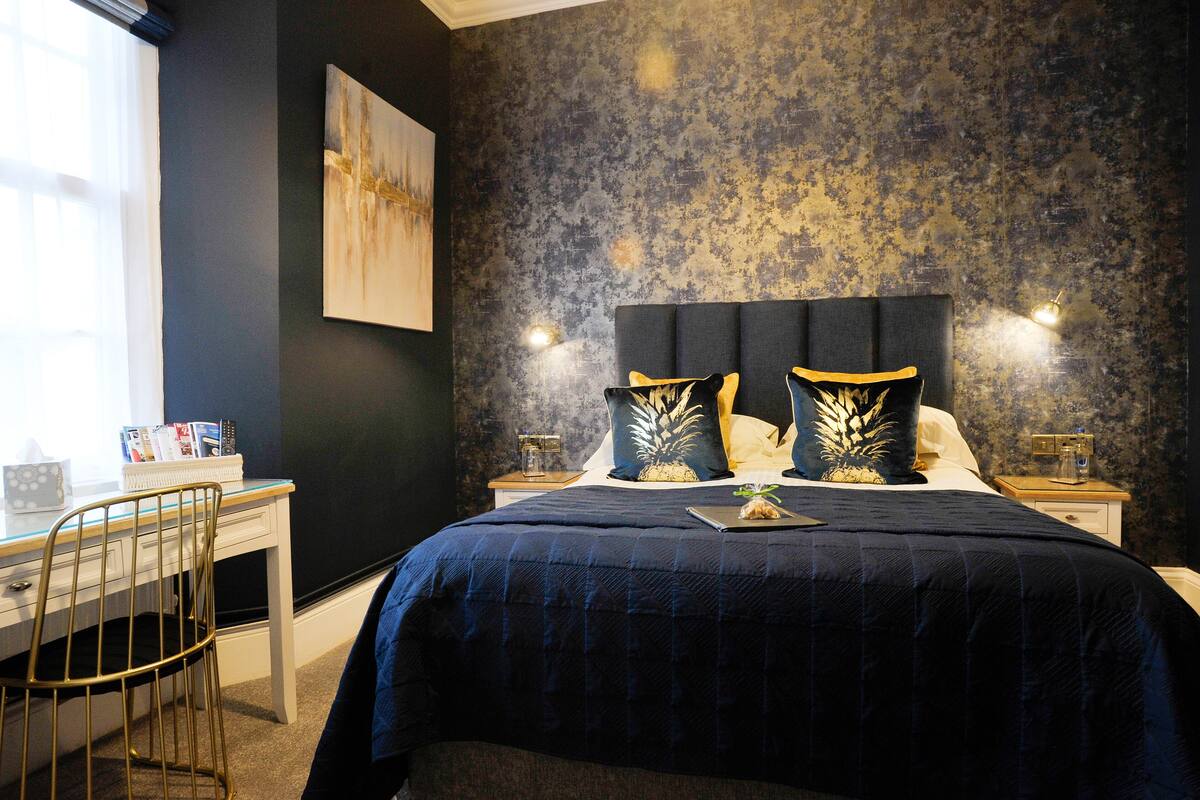 A bedroom with a dark blue bedspread, a headboard with two decorative pillows, a bedside table with a lamp, and a desk with a chair.