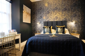 Quarto com colcha azul escura, cabeceira com dois travesseiros decorativos, mesa de cabeceira com abajur e escrivaninha com cadeira.