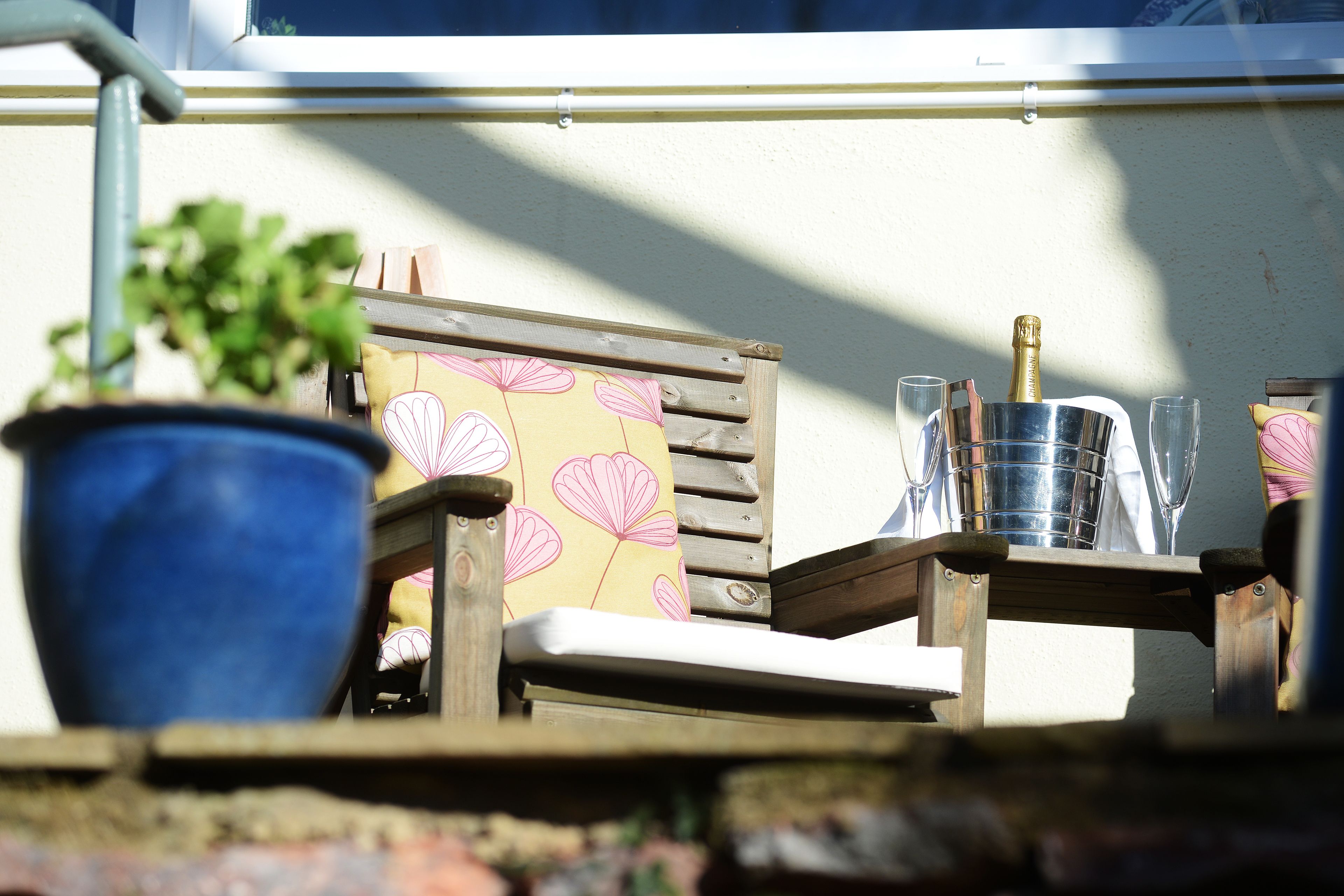 A wooden chair with a floral cushion, a potted plant, a metal bucket, and a bottle of champagne.
