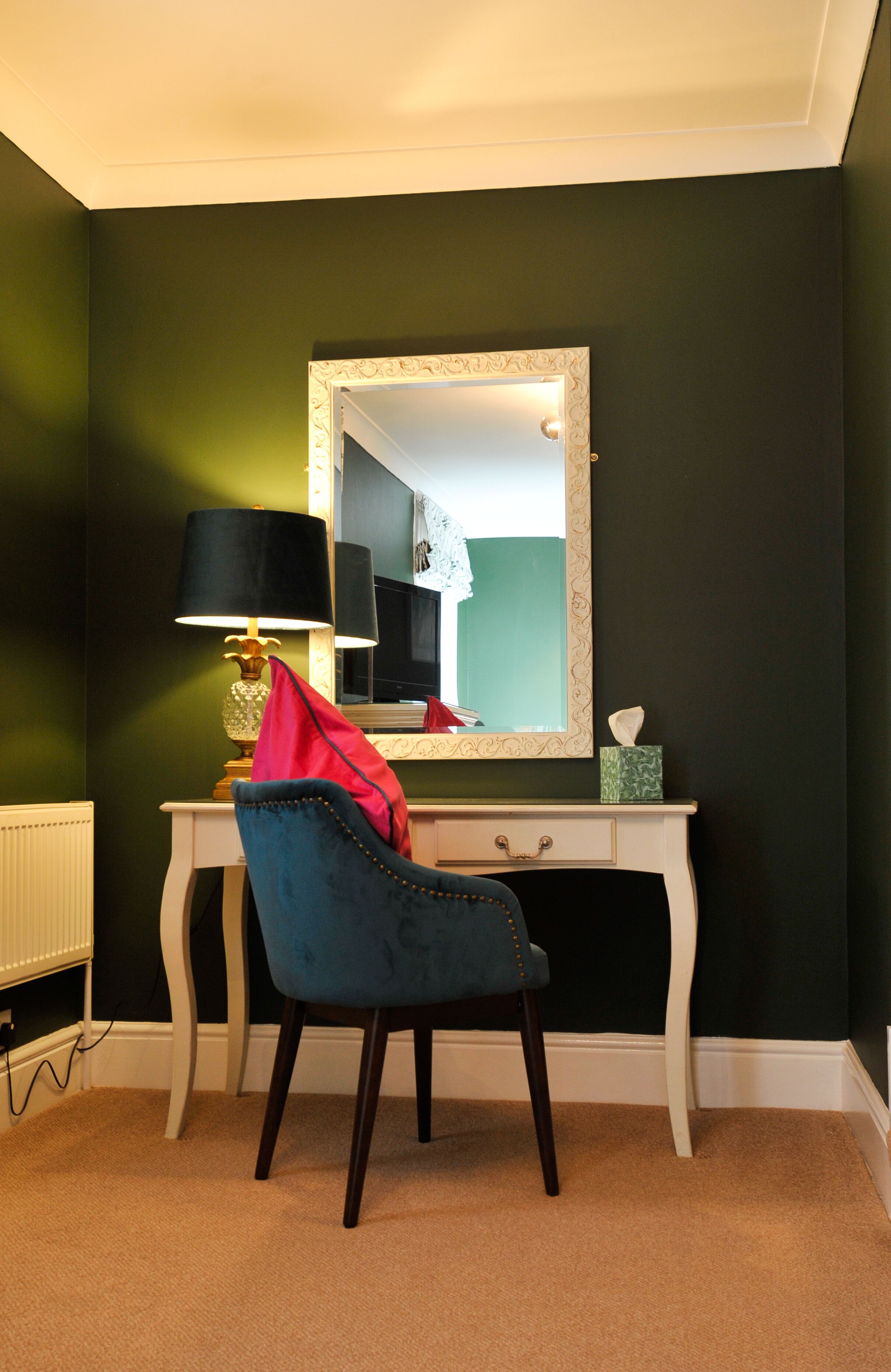 Una habitación con una pared verde oscuro, un tocador blanco con espejo, una silla azul, una lámpara y un radiador.