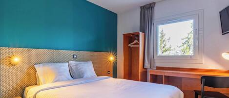 A hotel room with a large bed, a desk, a chair, a wardrobe, and a window with a view of trees.