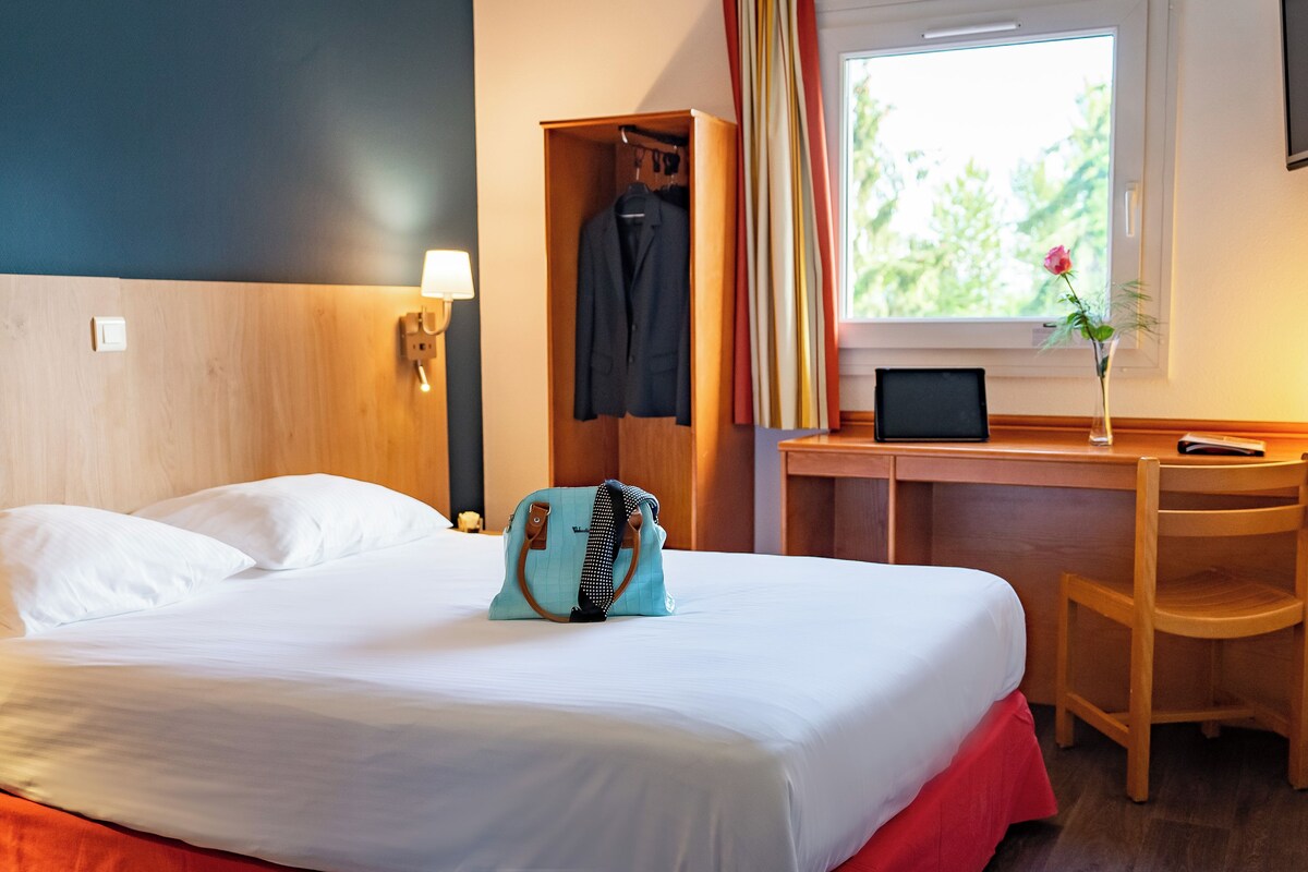 A hotel room with a bed, a desk with a chair, a laptop, a phone, and a window with curtains.