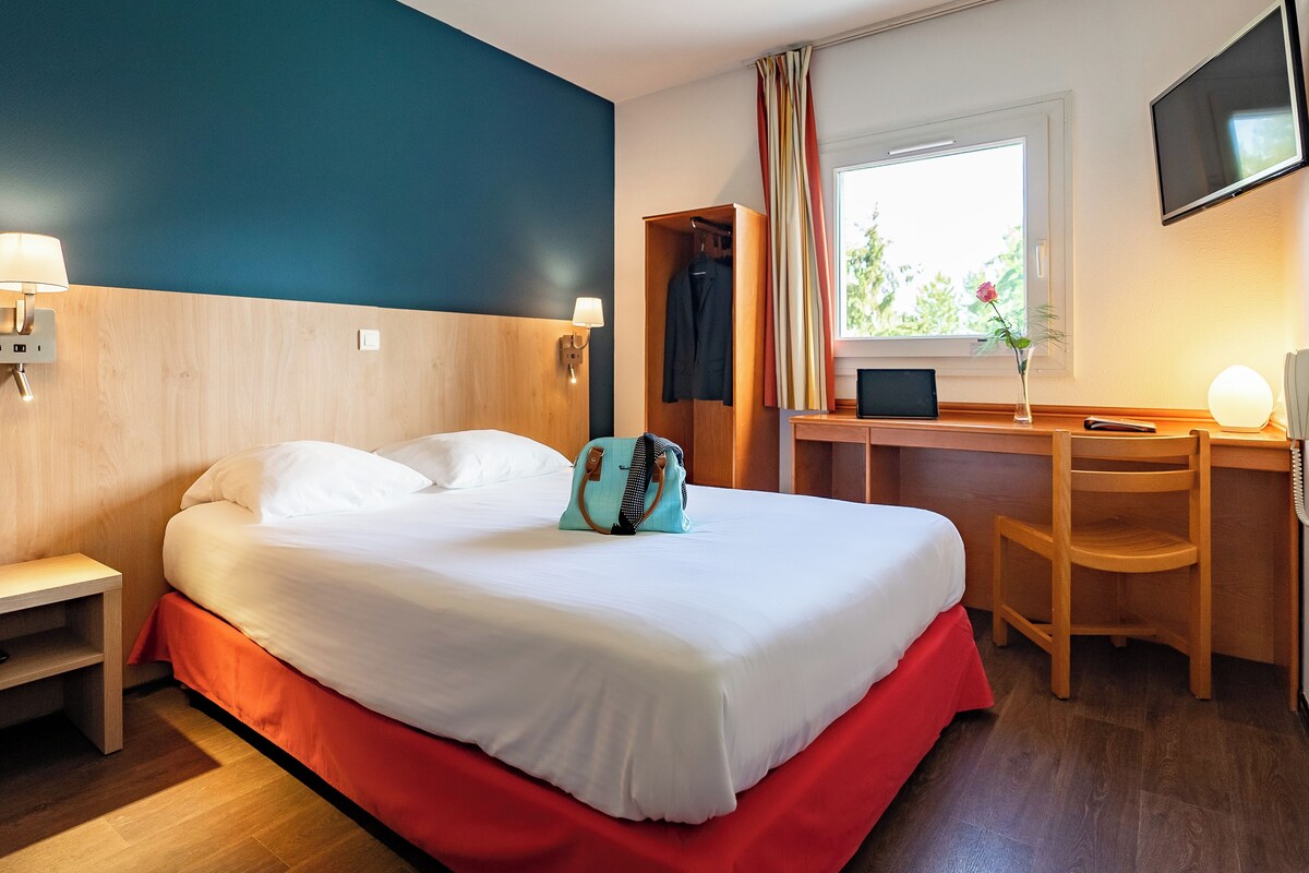 Une chambre d’hôtel comprenant un lit, une table de chevet, un bureau, une chaise et une fenêtre avec des rideaux.