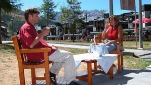 Dos personas sentadas a una mesa de madera con mantel blanco, al aire libre y con montañas al fondo.