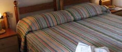 Habitación de hotel con cama doble, ropa de cama a rayas, cabecera de madera, mesita de noche con lámpara y velador con lámpara.