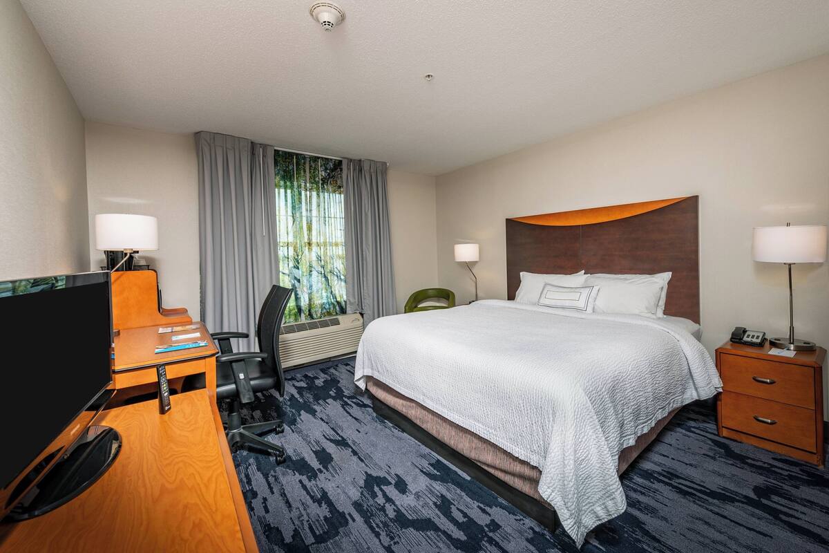 Habitación de hotel con cama, escritorio, silla, televisión y ventana con cortinas.