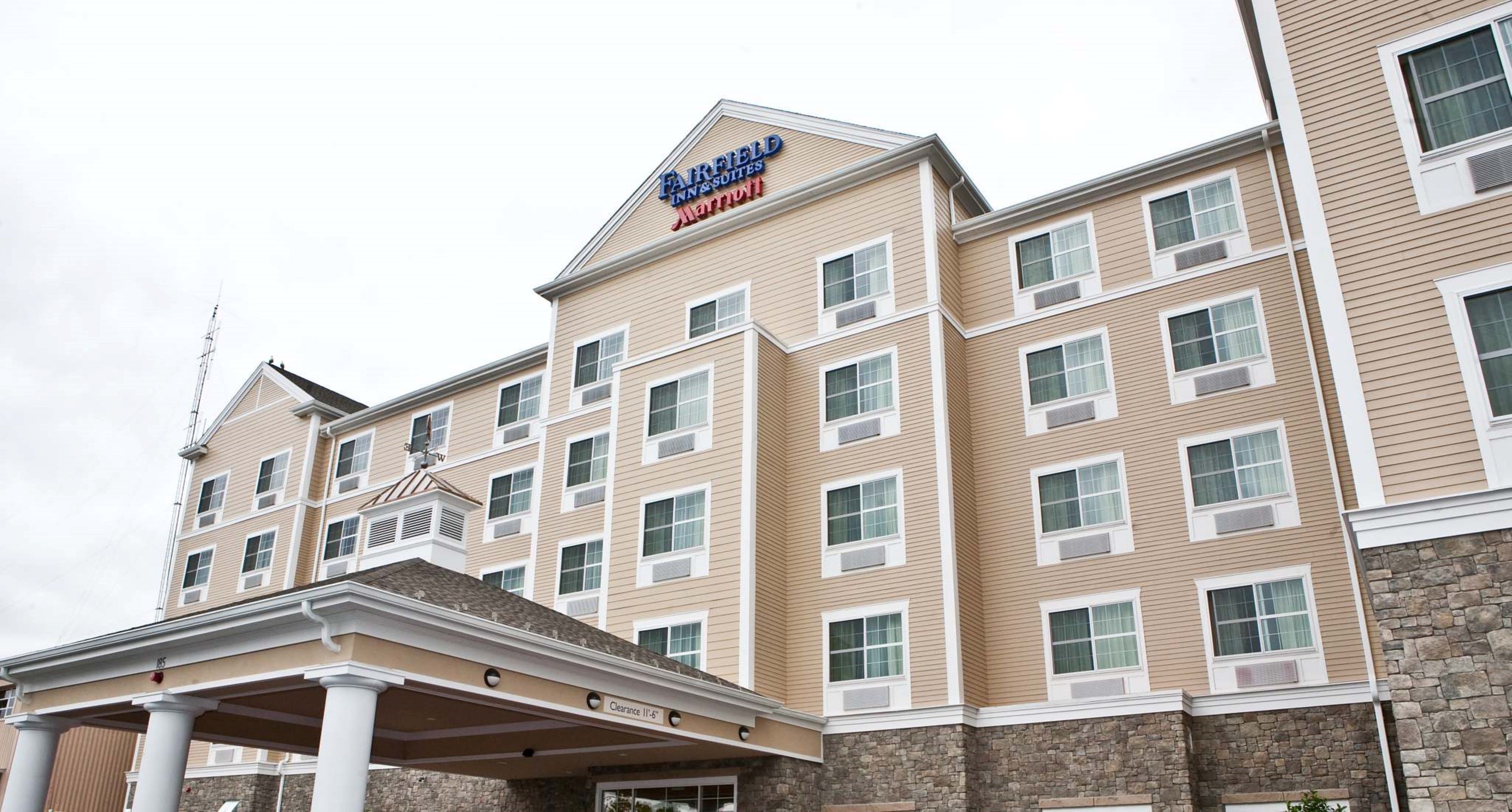 Un hotel de varios pisos con fachada color beige, una señal clara de Fairfield Inn & Suites by Marriott, y una entrada cubierta con columnas.